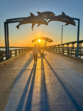 A person walks under a dolphin-themed arch on a pier in St. Augustine, Florida, at sunset, casting long shadows on the concrete walkway.