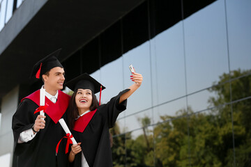 Couple of graduating students with diplomas taking selfie near university