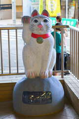 Tokyo, Japan - Sep 30 2024, vertical view of the sculpture of the white cat Maneki Neko on the street near the entrance to the subway station, with people on background, at daytime, Tokyo, Japan