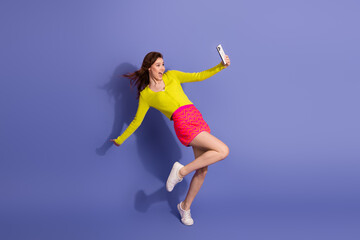 Energetic young woman posing playfully with smartphone against a vibrant purple background,...