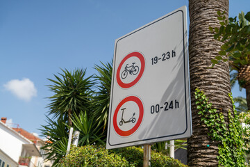 Sign showing time restrictions for bicycles and scooters, set against a vibrant tropical backdrop, featuring lush palm trees and clear blue sky.