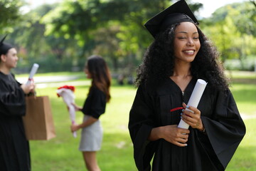 A graduate in cap and gown gazes upward with a joyful smile, holding her diploma. Behind her, two...