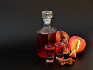Homemade apple-pomegranate liqueur on a black background, a bottle and two glasses of alcohol, cinnamon and anise nearby.