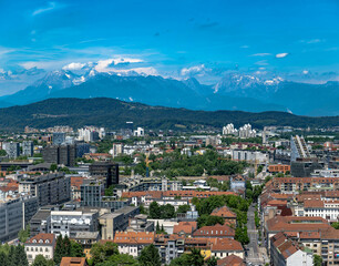Scenic Ljubljana cityscape featuring vibrant rooftops, lush greenery, and distant snowy mountains under a bright blue sky on a clear summer day