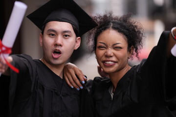 Two graduates in caps and gowns beam with joy, embracing and cheering in celebration. Their wide...