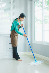 Asian housewife, dressed in a green sweater and brown apron, smiles while mopping the shiny floor near a sunlit window. Her dedicated cleaning keeps the home fresh, hygienic, and welcoming.