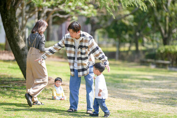 On a sunny March day in a lush forested park in Japan, a six-month-old Japanese baby boy sits on the grassy lawn, smiling happily while his thirtysomething mom and dad joyfully play with him nearby.