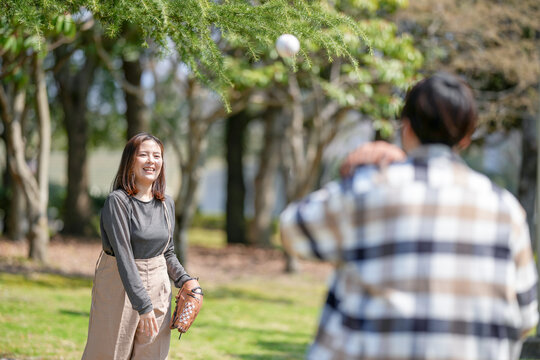 On a sunny March day in a forested park in Japan, a Japanese couple in their thirties, each wearing a baseball glove, play catch together, smiling as they toss the ball under the clear spring sky.