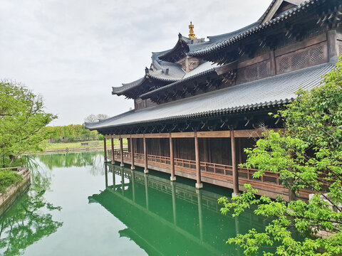 A serene temple complex with a pagoda reflects on a calm green lake, surrounded by lush trees and traditional architecture 
