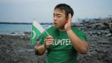 Young man on beach holding pakistan flag and cupping ear as if listening intently, wearing green volunteer shirt, representing cultural diversity by the sea. - Powered by Adobe