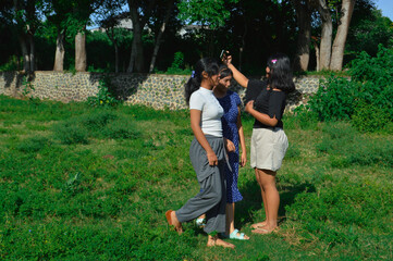 Fototapeta premium One girl holds up her phone to take a selfie while the other two walk toward her across the grassy field.