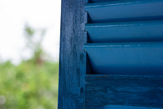 Close-up of a blue wooden louvered window with rustic texture, blurred green foliage background. Architectural detail, vintage style home decor - Powered by Adobe
