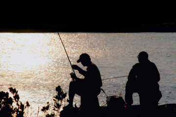 Two fishermen silhouetted against the sparkling water at sunset in Houlong Port, Miaoli County, Taiwan. The scene captures the tranquility of an evening fishing trip.