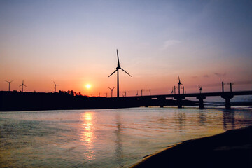 Silhouetted windmills and a bridge are visible against a vibrant sunset over the water in Houlong Port, Miaoli County, Taiwan. Capturing the beauty of renewable energy in coastal landscapes.