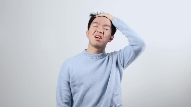 A young Asian man in a light blue sweater performs a facepalm gesture, closing his eyes and pressing a hand to his face in exasperation. Conveying regret, embarrassment, or disbelief.