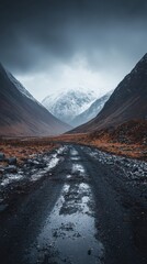 Remote mountain landscape with rugged dirt road leading through deep valleys, snow capped peaks under dramatic cloudy sky, moody wilderness scene