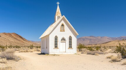 Small white wooden church with steeple and cross situated in a desert landscape under clear blue sky with distant mountains and sparse vegetation
