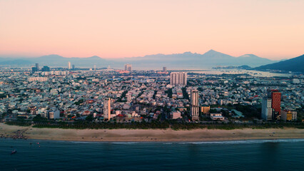 Aerial panorama revealing da nang skyline glowing at sunset, modern architecture framing sandy coastline, serene ocean waters, mountainous silhouettes defining vietnamese landscape
