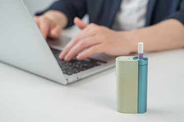 Business woman smoking an electronic cigarette while working on a laptop. Tobacco heating system in...