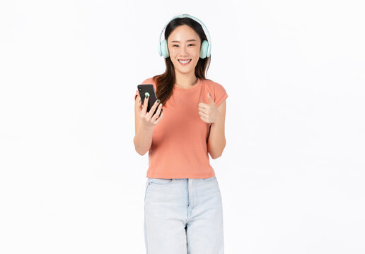 Cheerful young Asian woman in headphones listening to music and enjoy favourite playlist application on smartphone with dancing on white background.