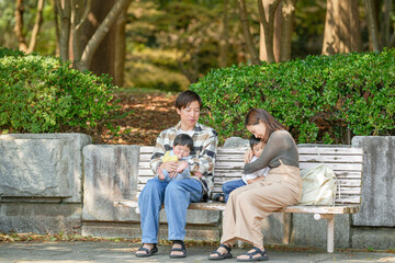 In March, at a sunny park in Fukuoka, a Japanese couple in their thirties sits on a bench with their 3-year-old son and baby girl, 6 months old. This photo captures a peaceful family moment outdoors.