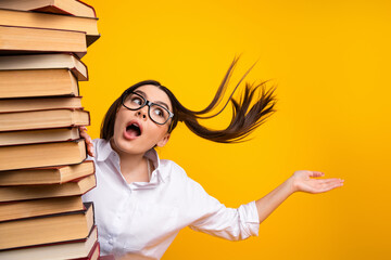 Excited young student woman in white shirt posing beside a tall stack of book on vibrant yellow...