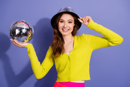 Smiling young woman in vibrant attire holding a disco ball against a purple background expressing joy and style