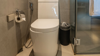 A white toilet in the corner of the bathroom. The urn is nearby. Hygienic shower. There is a roll of toilet paper on the beige tiled wall. The shower cabin is behind a glass partition. Towel.