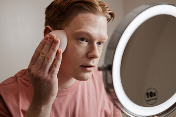 A Close-up Of A Young Man Applying Foundation Skincare Makeup Routine
A young man applies foundation using a makeup sponge while standing in front of a mirror with an LED ring light. 