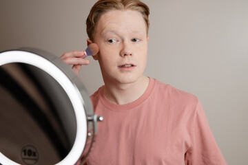 A Close-up of A Young Man Applying Blush In Front Of A Ring Light Mirror
Young man applying makeup with a brush in front of a lighted vanity mirror. 