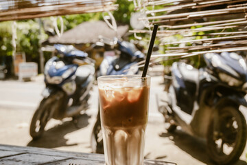 Refreshing iced coffee latte served in a glass with straw outdoor at daytime on the terrace of a restaurant in General Luna on Siargao in the Philippines with blurry background.
