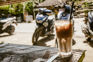 Refreshing iced coffee latte served in a glass with straw outdoor at daytime on the terrace of a restaurant in General Luna on Siargao in the Philippines with blurry background.