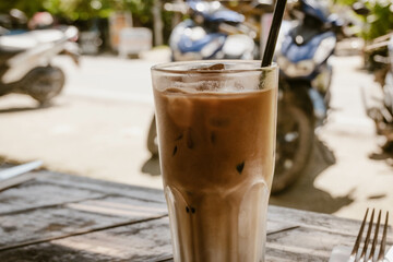 Refreshing iced coffee latte served in a glass with straw outdoor at daytime on the terrace of a restaurant in General Luna on Siargao in the Philippines with blurry background.