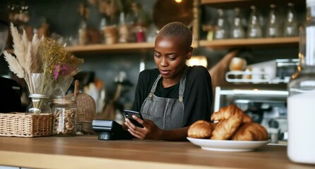 Young african barista uses smartphone at the counter of a coffee shop with croissants in the foreground - Powered by Adobe