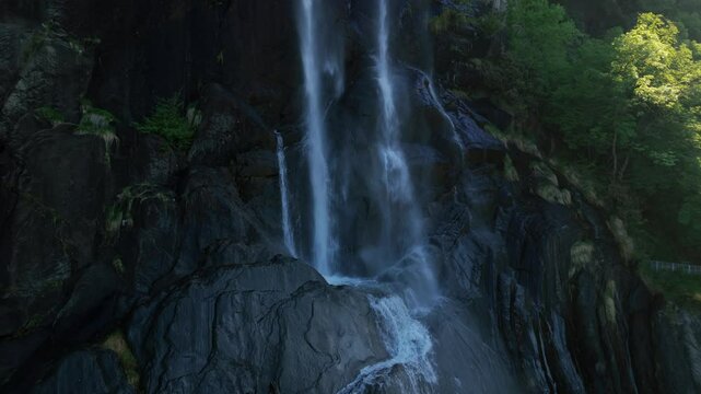 Acquafraggia waterfalls, water flowing over dark rocks, lush sunlit green trees, Val Bregaglia, Lombardy, Italy. Aerial drone, close-up