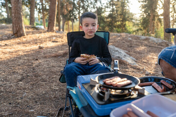 A young boy sits in a camping chair, holding a sandwich while hot dogs sizzle on a small grill. The sun sets behind tall trees, creating a peaceful camping atmosphere.