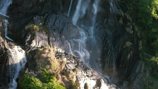Acquafraggia waterfalls cascading down rocky cliffs, lush greenery, sunlight, Val Bregaglia, Lombardy, Italy. Aerial drone backward