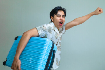 Happy young Asian man in summer shirt shouting "GO" while carrying a bright blue suitcase, posing with excitement against a plain background, ready for a fun holiday or travel adventure