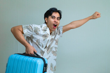 Happy young Asian man in summer shirt shouting "GO" while carrying a bright blue suitcase, posing with excitement against a plain background, ready for a fun holiday or travel adventure