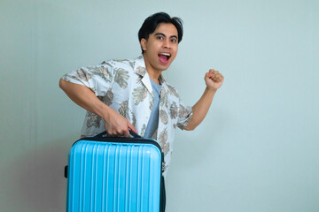 Excited young Asian man in summer clothes posing with a running gesture while holding a blue suitcase, isolated on a clear background, ready for vacation or travel adventure