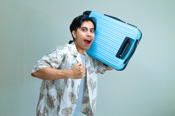 Excited young Asian man in summer clothes posing with a running gesture while holding a blue suitcase, isolated on a clear background, ready for vacation or travel adventure