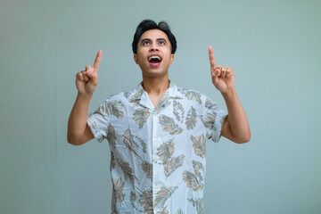 Happy young Asian man in summer clothes smiling and pointing up with both hands against a clear background, showing excitement and positivity, perfect for promotion or advertisement