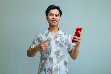 Portrait of a smiling young Asian man in summer clothes pointing at a cellphone he is holding, isolated on a clear background, perfect for digital ads, promotions, or mobile app concepts