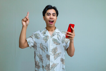 Excited young Asian man in summer clothes pointing up while holding a cellphone, standing against a clear background, showing cheerful mood and vibrant holiday energy