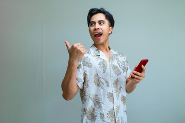 Happy young Asian man in summer clothes pointing aside with his thumb while holding a smartphone, isolated on a clear background, showing excitement and joy