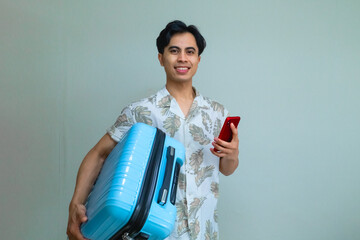 Happy young Asian man in summer outfit smiling while using smartphone and carrying a blue suitcase, ready for travel or vacation, isolated on a clear background