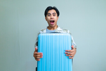 Happy young Asian man in summer clothes hugging a suitcase with a wow expression, looking at the camera, isolated on a clear background, ready for vacation or travel