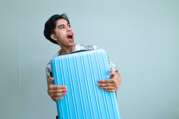 Portrait of a young Asian man in summer outfit looking to the side with a surprised wow expression while hugging a suitcase, isolated on a clear background, travel concept