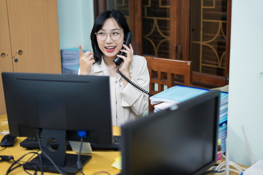 Young Asian woman communicating on phone while seated at office desk with computer and paperwork during a busy workday