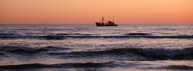 A lone fishing boat sails across calm waters as the sun sets behind the horizon, casting vibrant hues of orange and pink across the sky. Vejers Strand beach Denmark